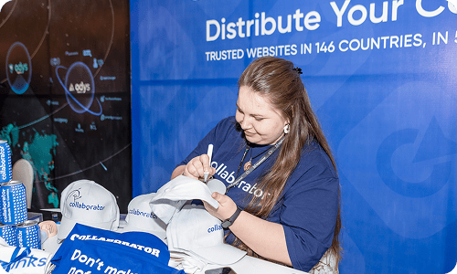 Collaborator team member in a branded shirt signing promotional t-shirts and materials at a booth at the SEO Mastery Summit, with Collaborator’s global content distribution messaging visible in the background